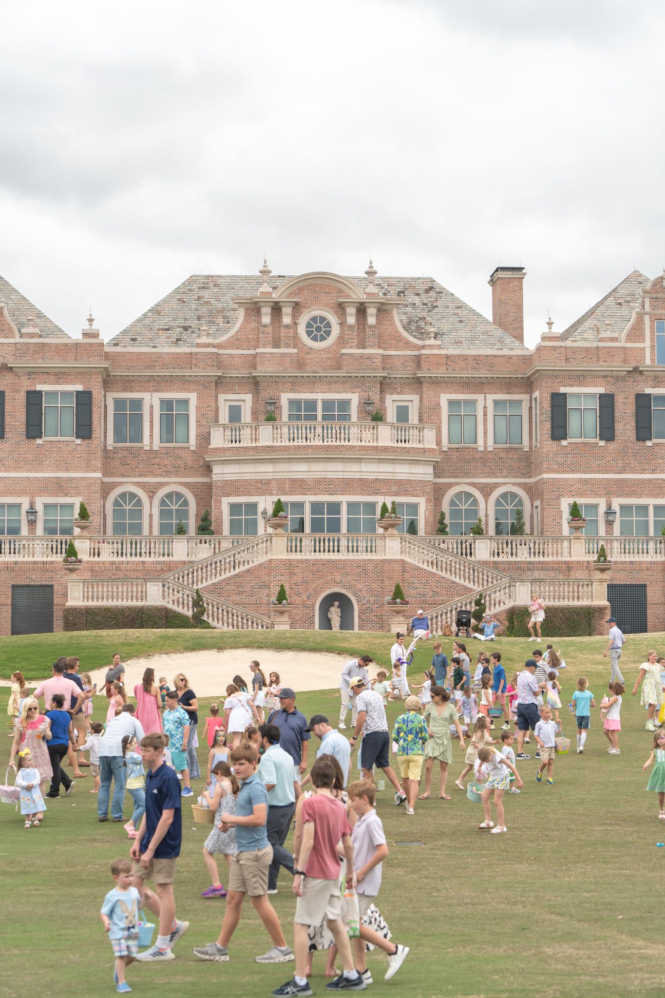 Large crowd of people spread across a lawn during an outdoor event in front of a grand brick mansion-style building.