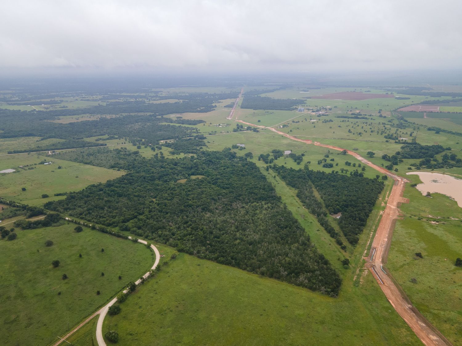 Aerial view of a rural landscape with green fields divided by dirt roads and forested areas stretching to the horizon.
