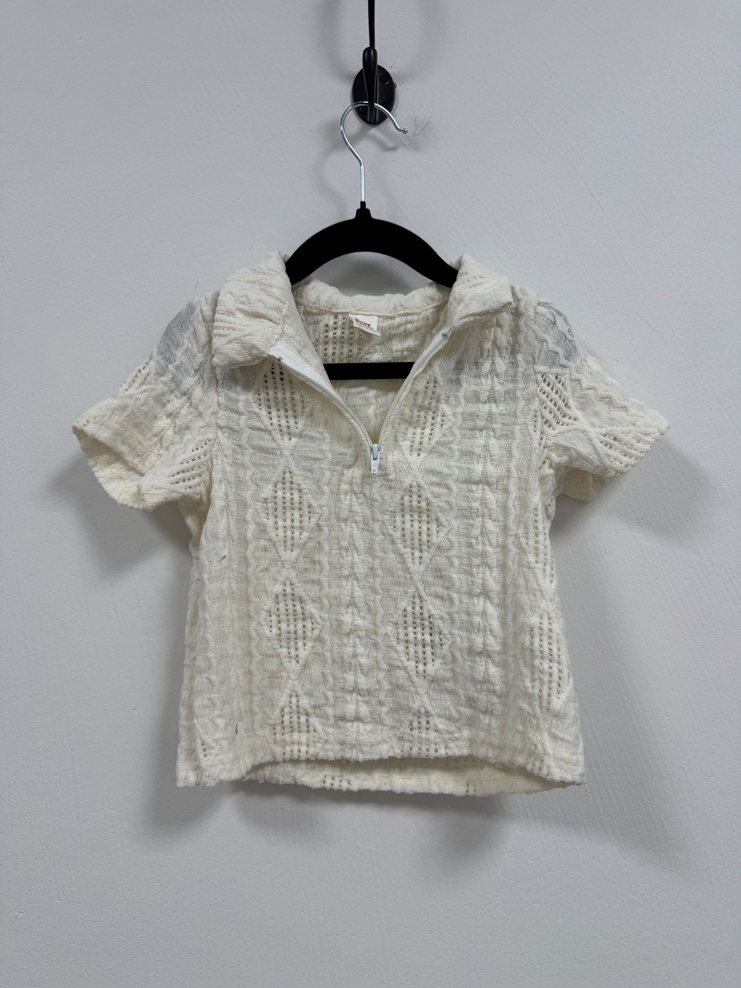 Cream colored short-sleeve textured blouse with collared neckline hanging on black hanger against white wall.