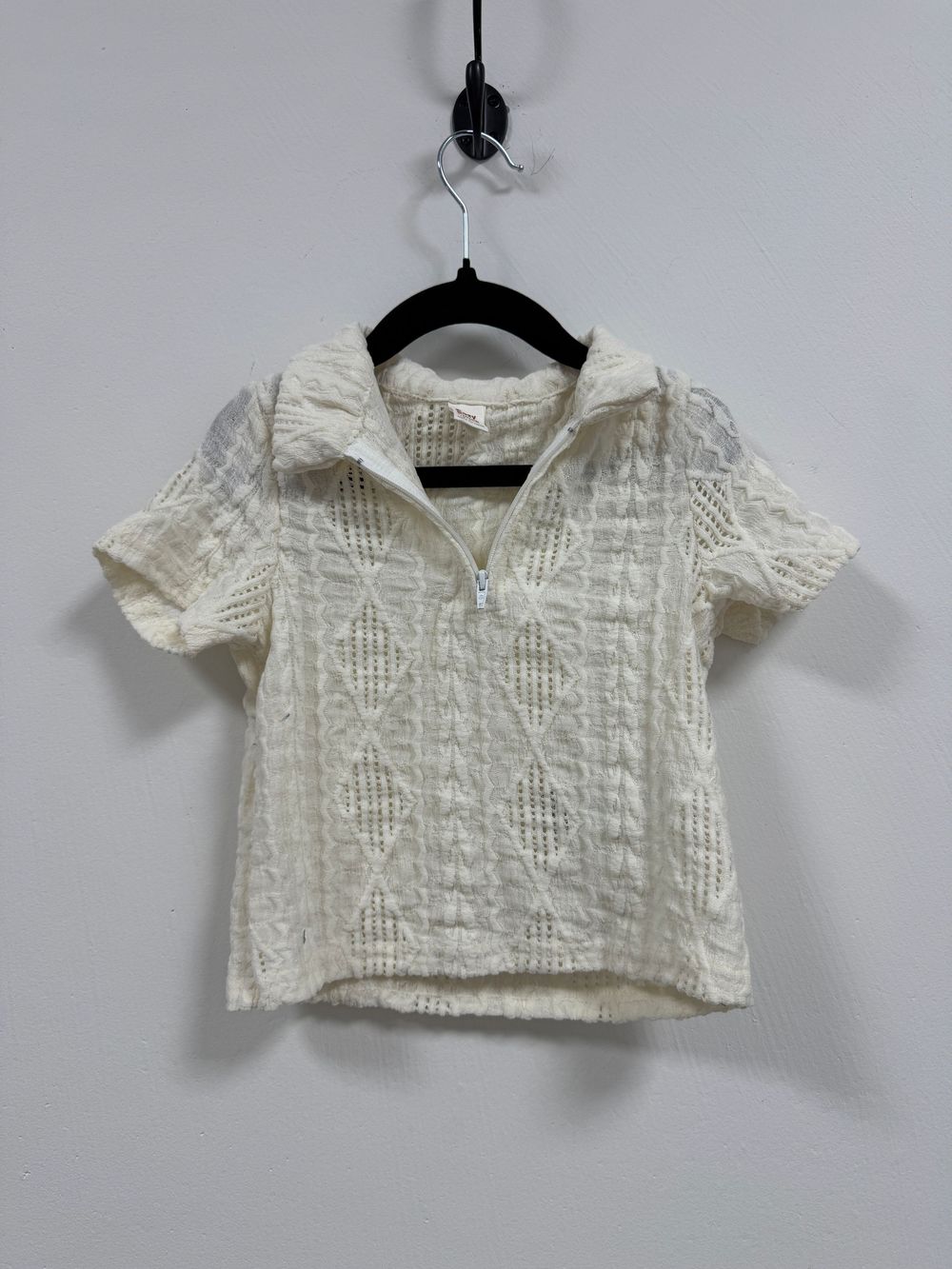 Cream colored short-sleeve textured blouse with collared neckline hanging on black hanger against white wall.