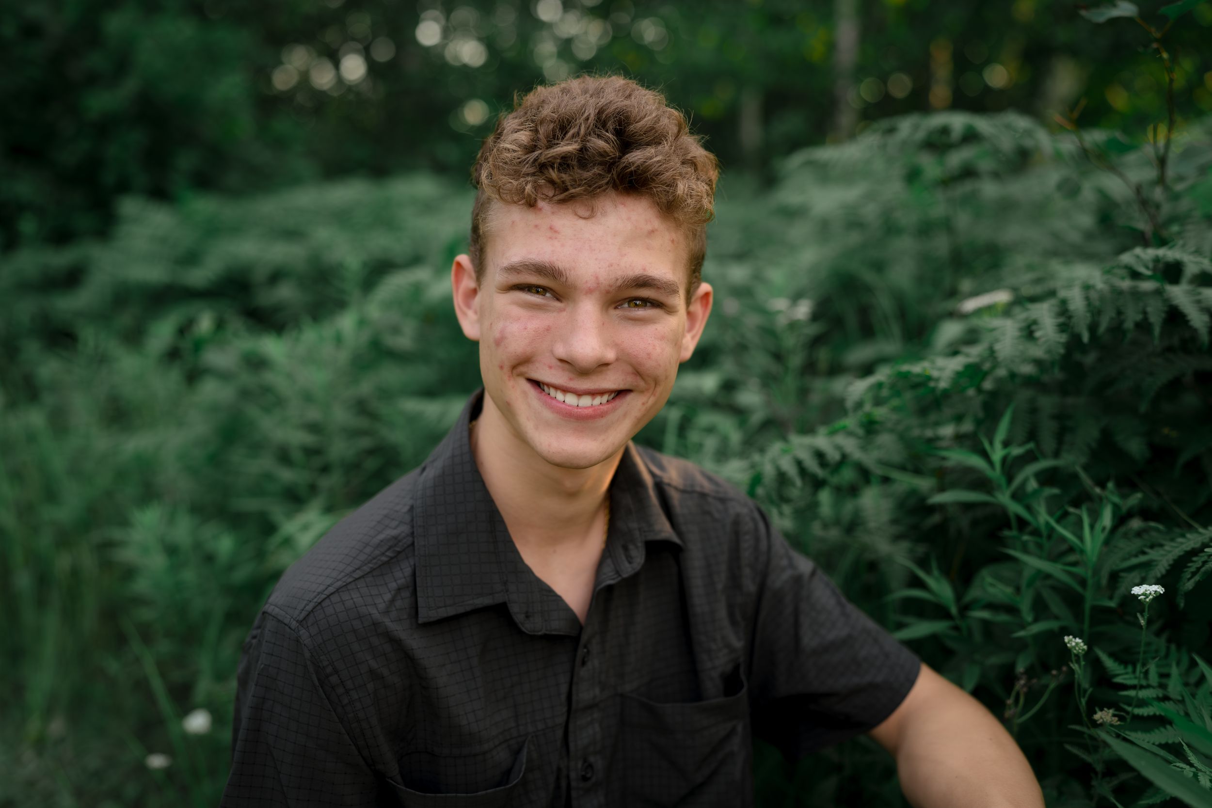 A person wearing a black shirt smiles warmly while sitting in front of lush green foliage outdoors.