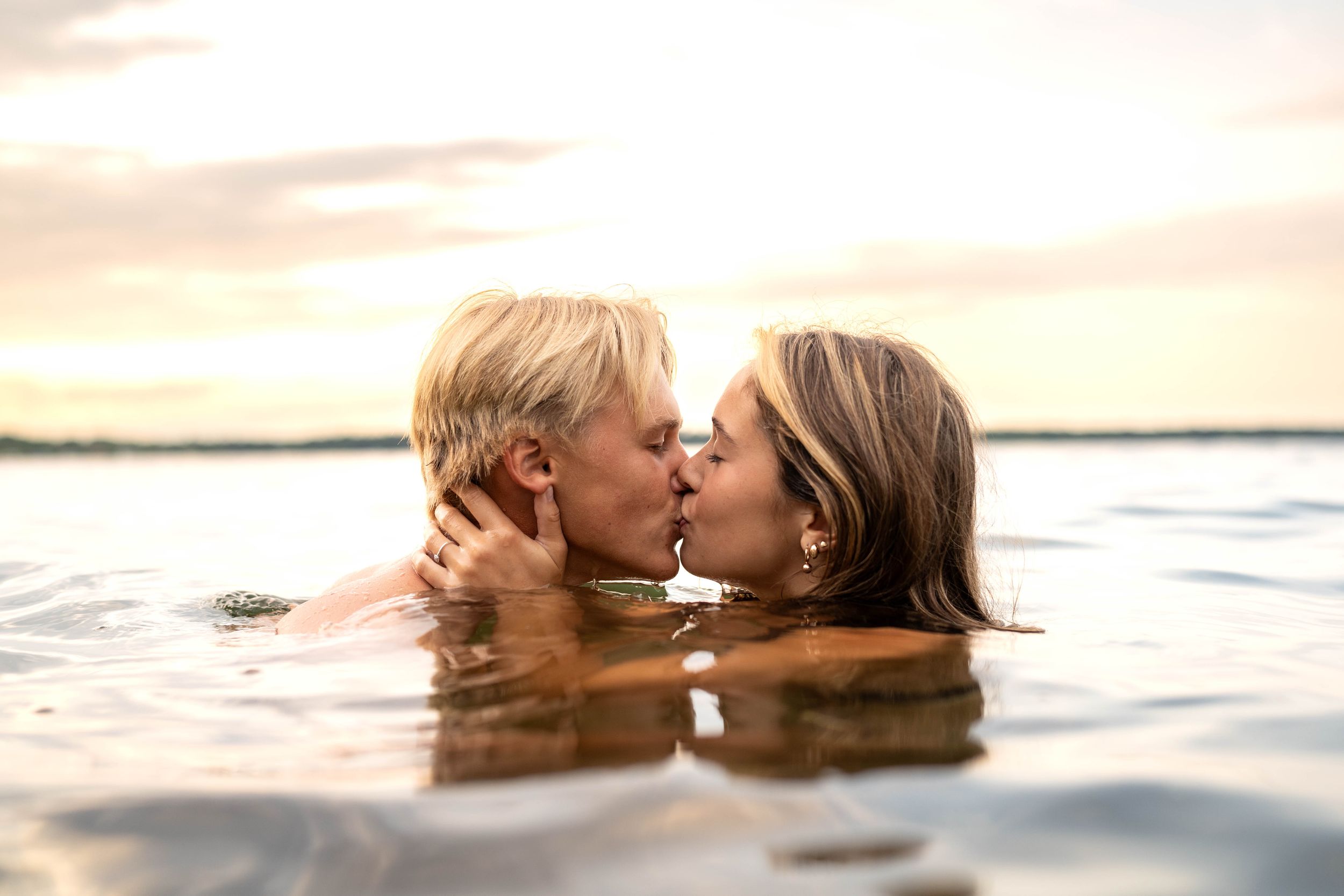 Two people share an intimate moment kissing while swimming in calm waters during a beautiful sunset.