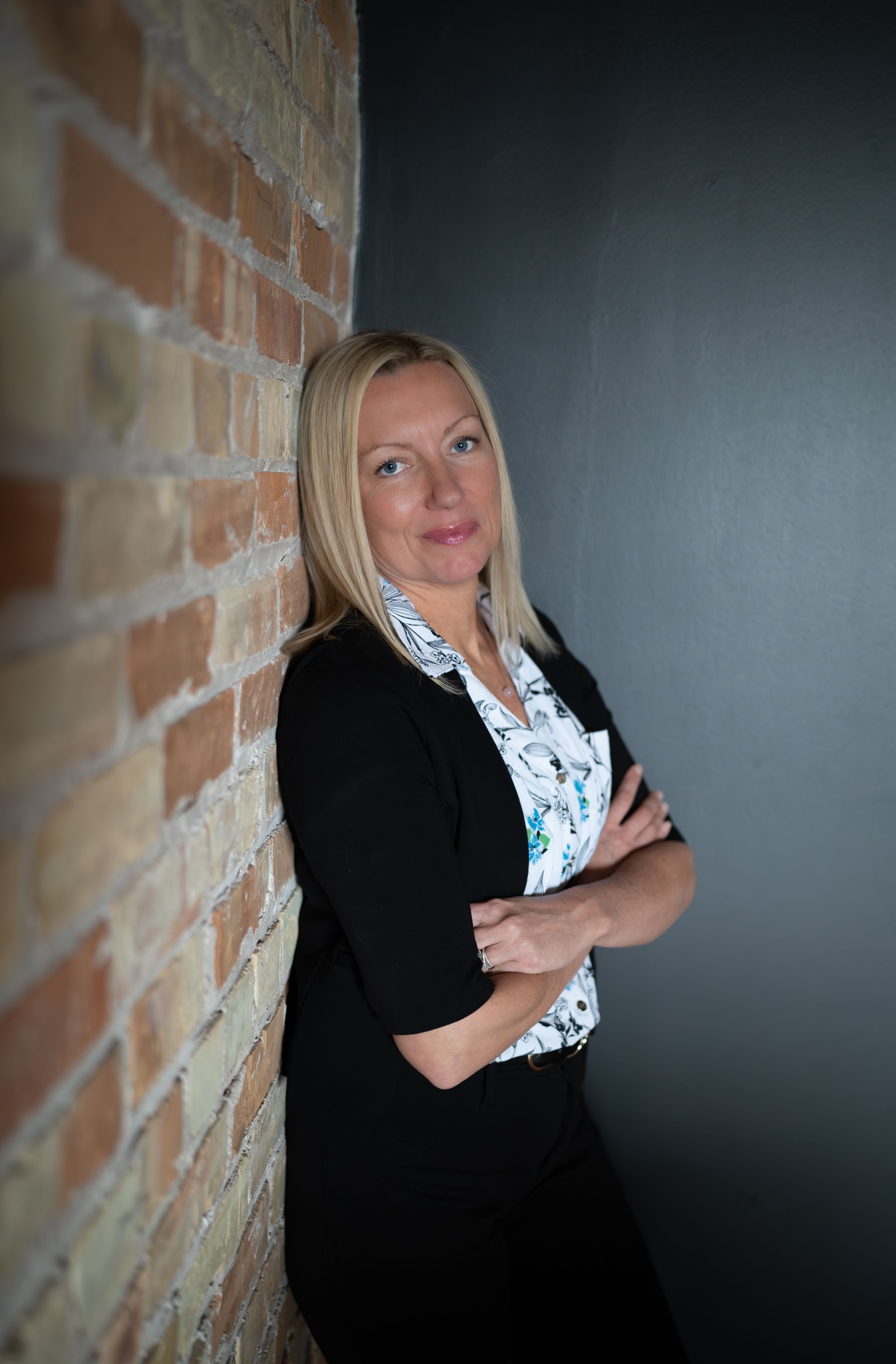 Professional portrait of someone in business attire leaning against a brick wall in moody lighting.
