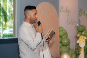 Man in white shirt speaking into microphone at a Baby Cortez baby shower celebration event.