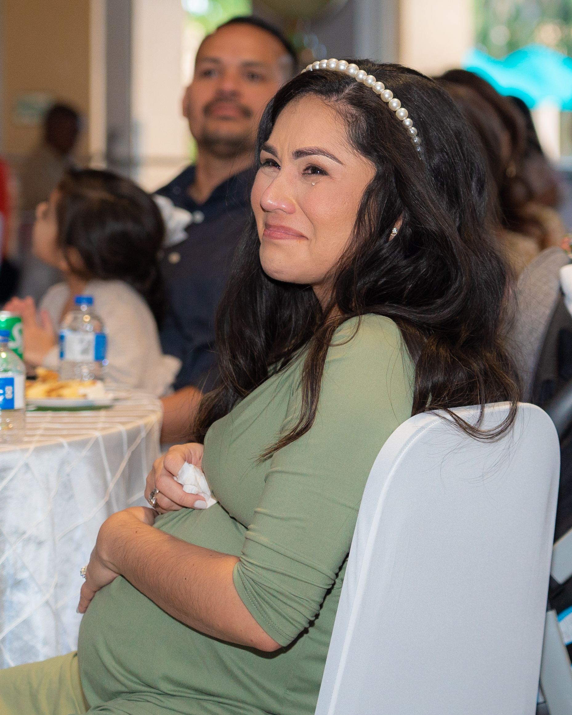 Pregnant woman in sage green dress smiling warmly while seated at a baby shower celebration.