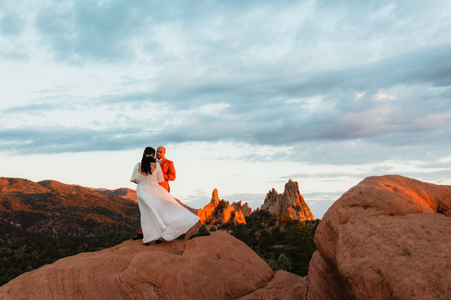 Silhouetted couple sits on desert cliff edge overlooking majestic red rock formations during golden sunset.