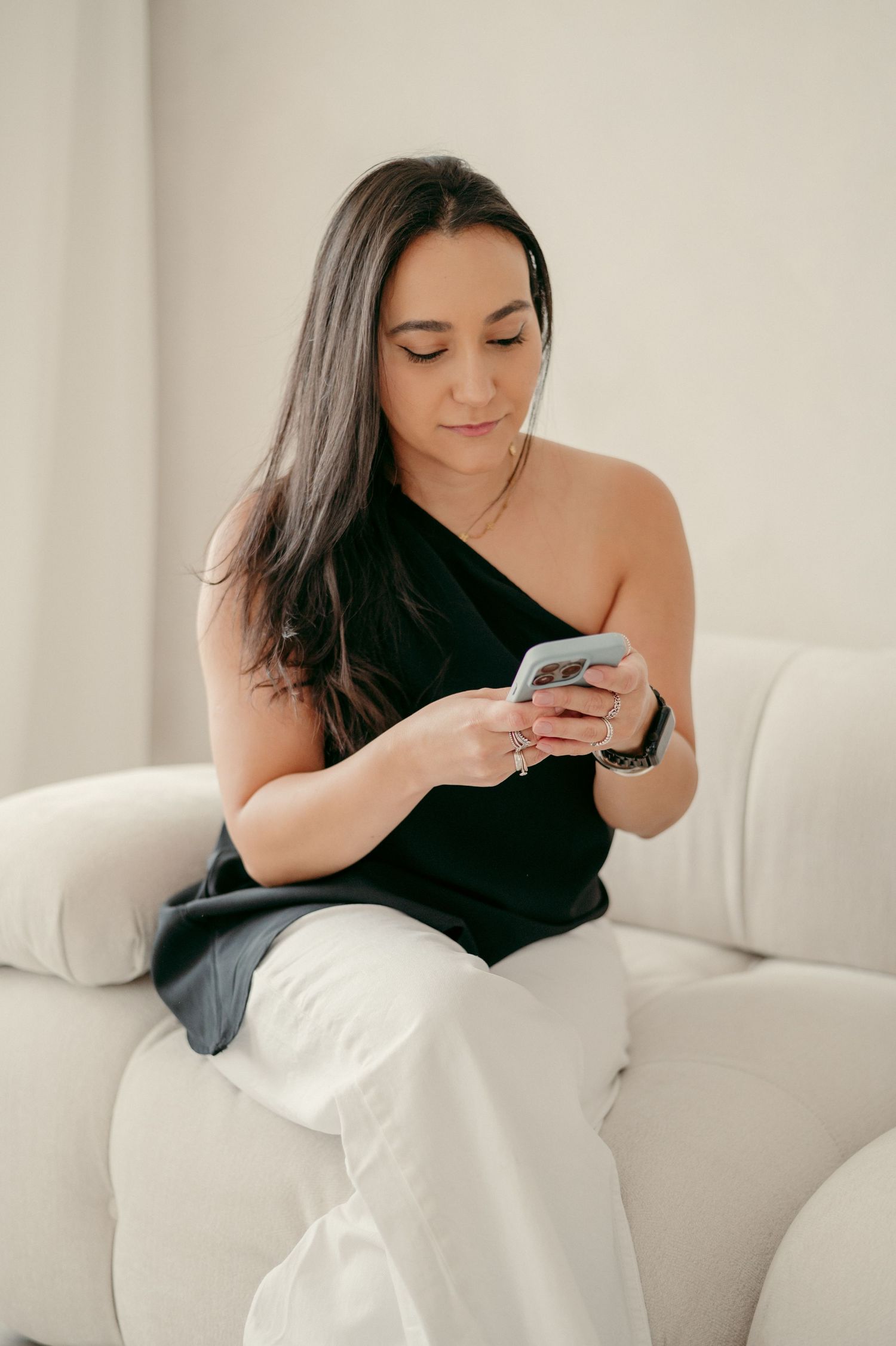 A person in a black one-shoulder top checks their phone while sitting on a white couch.