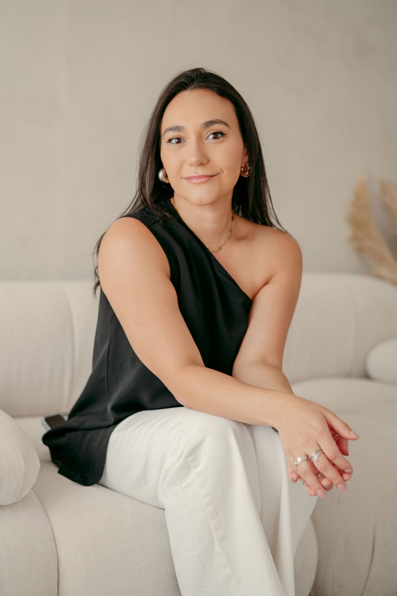 A person in an asymmetrical black top and white pants sits elegantly on a modern white couch.