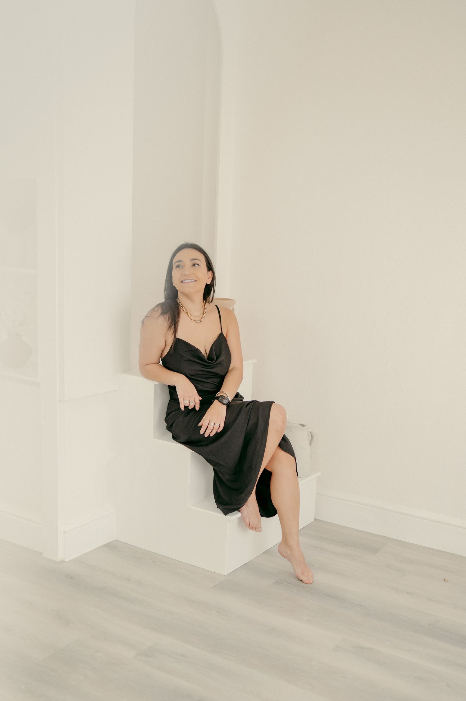 A person in a black dress sits barefoot on a white ledge in a minimalist interior setting.