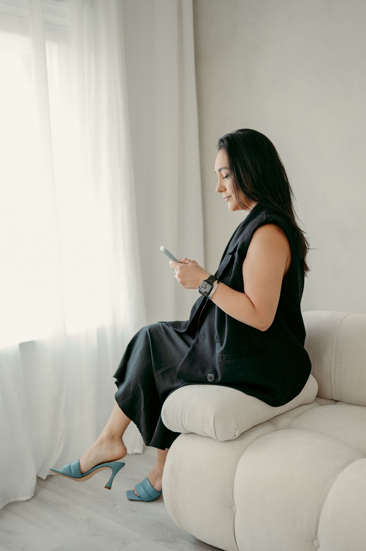A person in a black dress sits on a white couch checking their phone near sheer curtains.