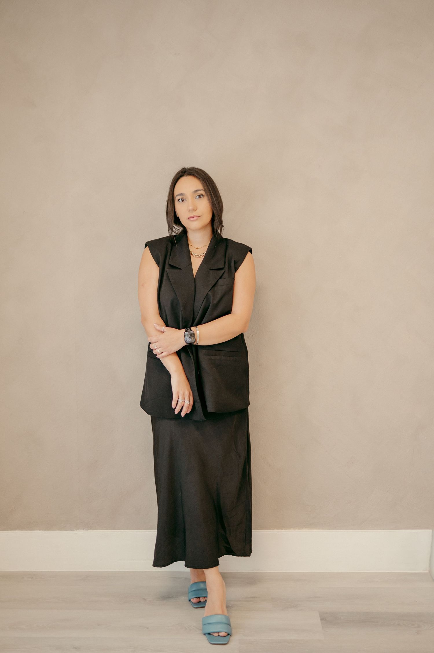 A person in a black ensemble and blue sandals poses against a neutral wall in a modern setting.