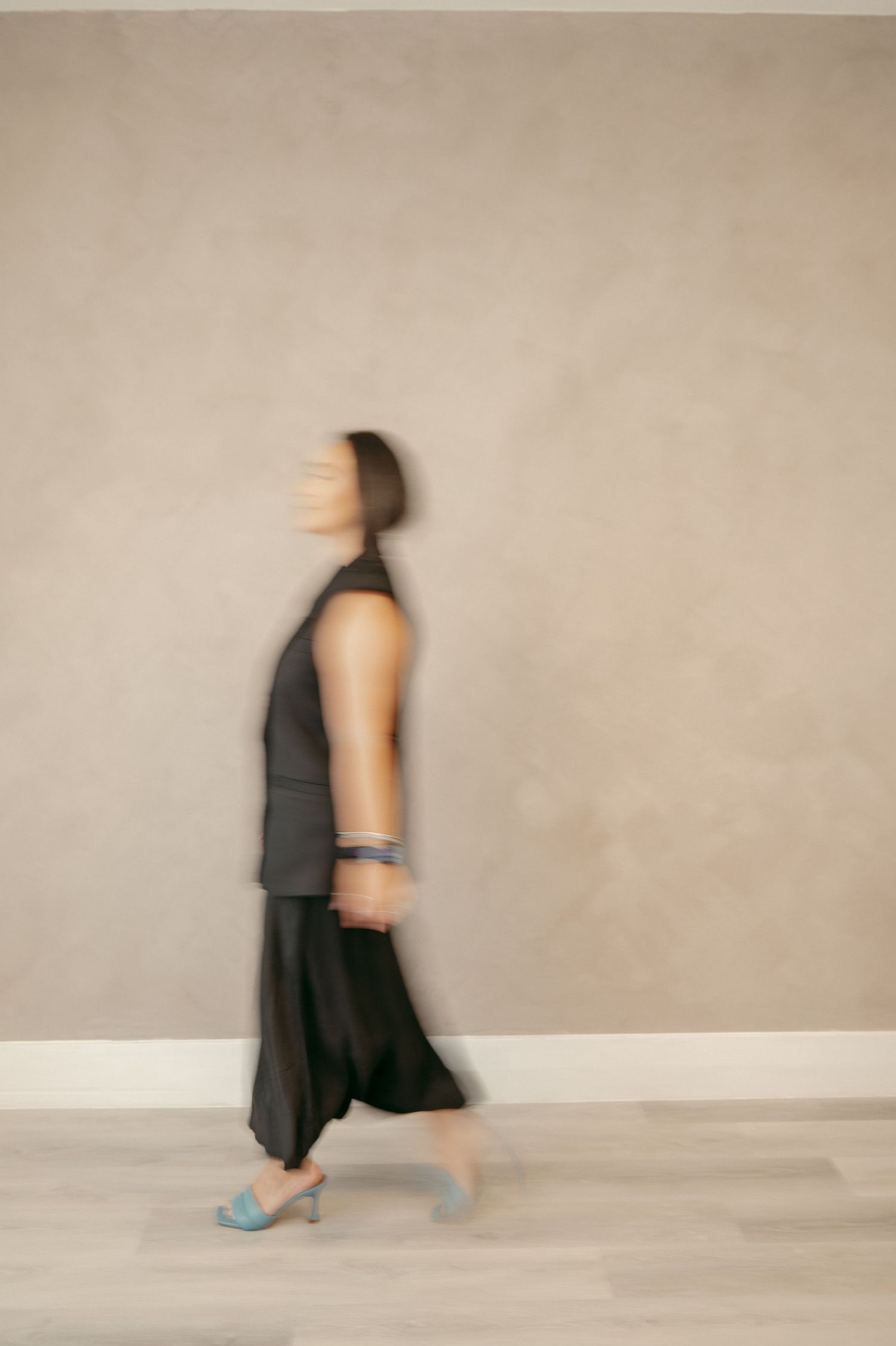 A motion-blurred figure in dark clothing walks past a beige wall wearing blue sandals.