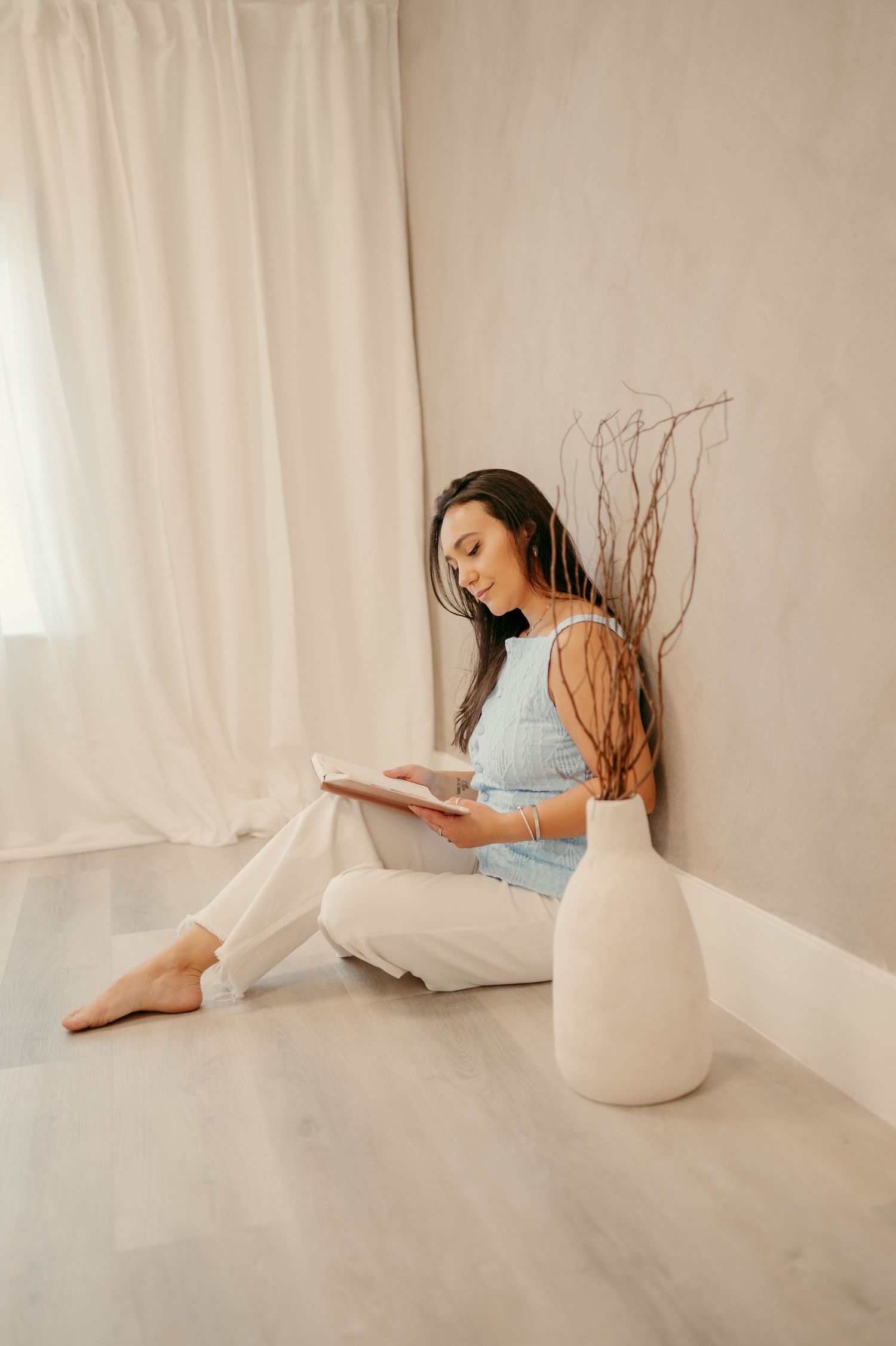 A person reads while sitting on the floor next to a white vase and decorative branches.