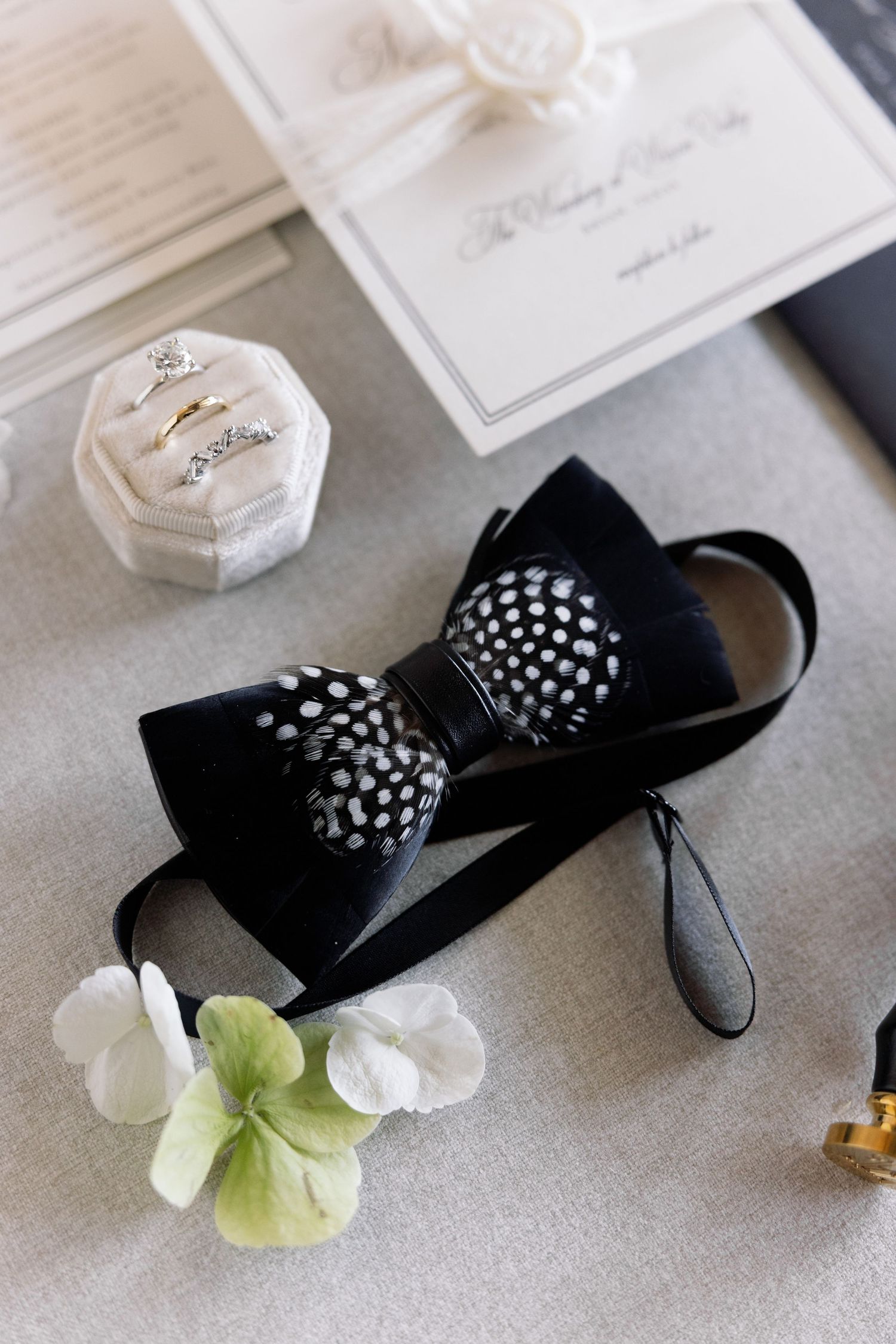 Wedding details featuring a black bow tie with polka dots, white ring box, and delicate flower petals on a light gray surface.