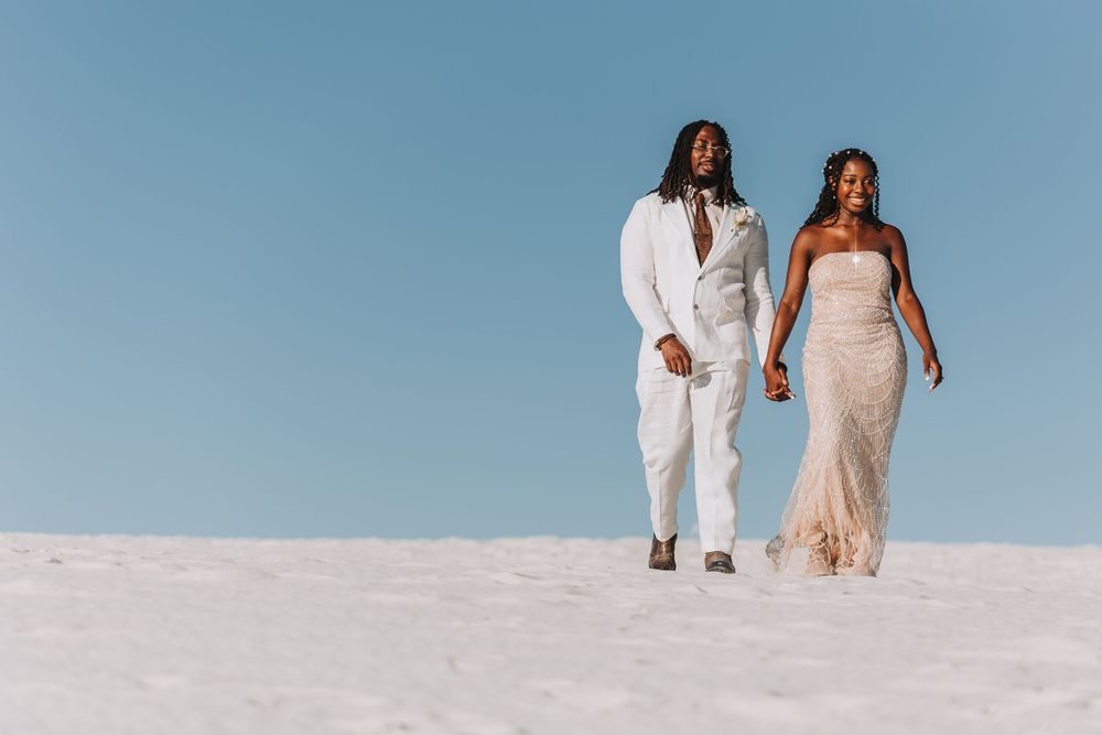 Two figures in white attire stand together on pristine white sand against a bright blue sky creating a romantic scene.