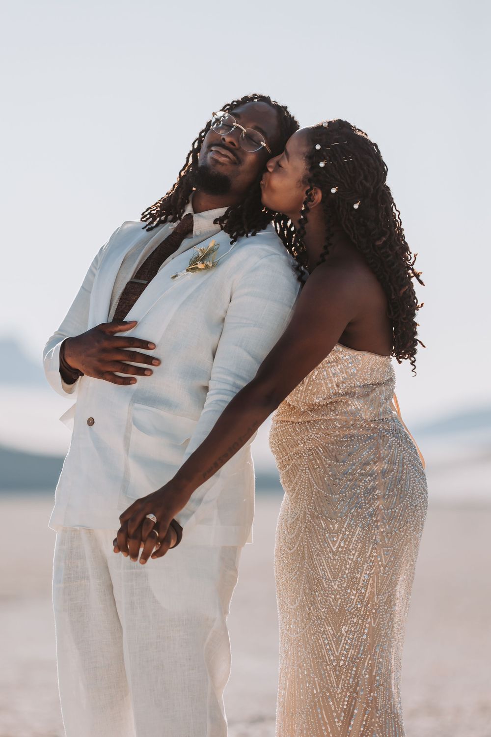 A loving couple share an intimate moment together on the beach, wearing elegant white and beaded formal attire.