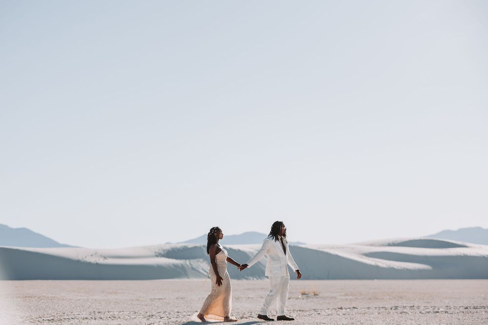 Two people in white outfits hold hands while walking across scenic white sand dunes under a pale blue sky.