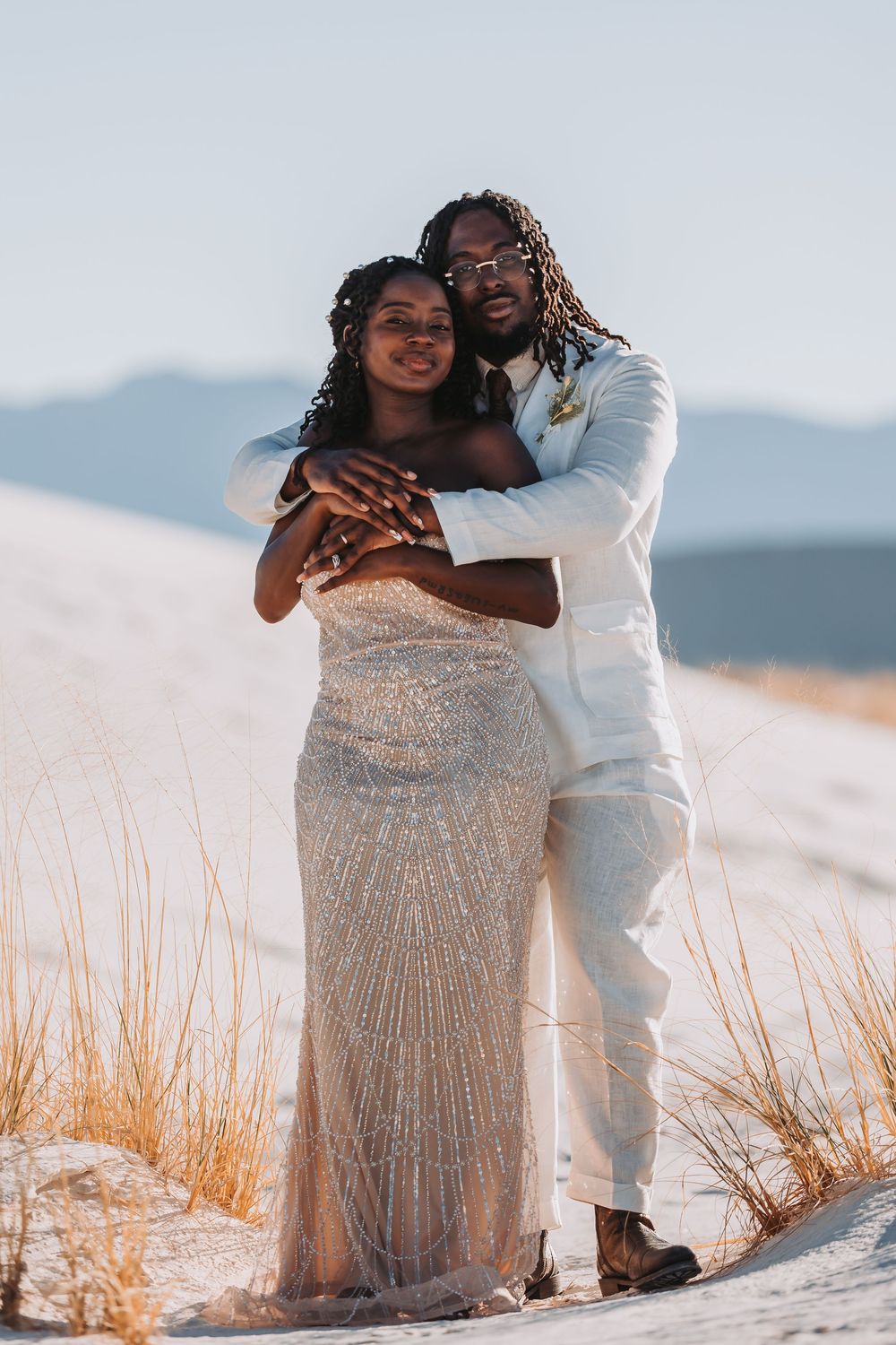 A couple embraces on sand dunes wearing elegant white outfits against a mountain backdrop at sunset.