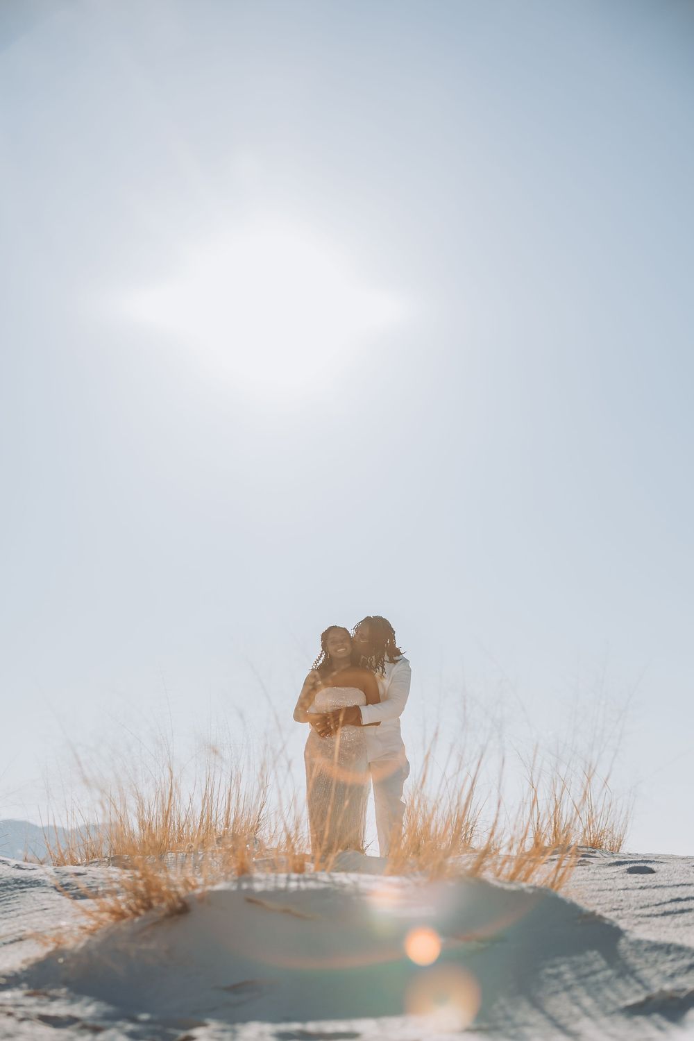 Two figures embrace on sandy dunes with tall grass and bright sunlight creating a dreamy backlit silhouette.