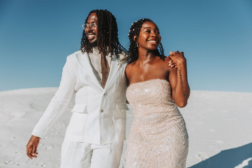 Happy couple poses playfully against a bright blue sky in coordinated white and beige wedding attire on their special day.