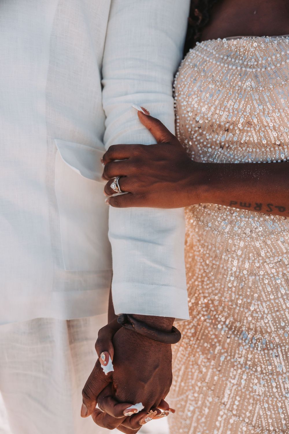 Close-up of couple holding hands showing wedding rings, with contrast between white suit and sparkly beaded dress.