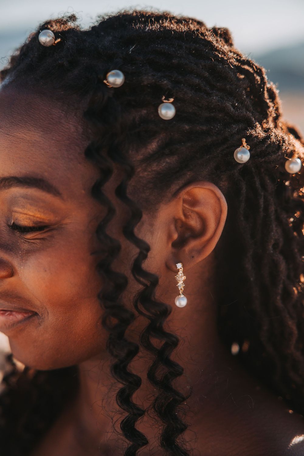 Close-up portrait of a wavy hairstyle adorned with pearl accessories and a delicate drop earring in warm sunlight.