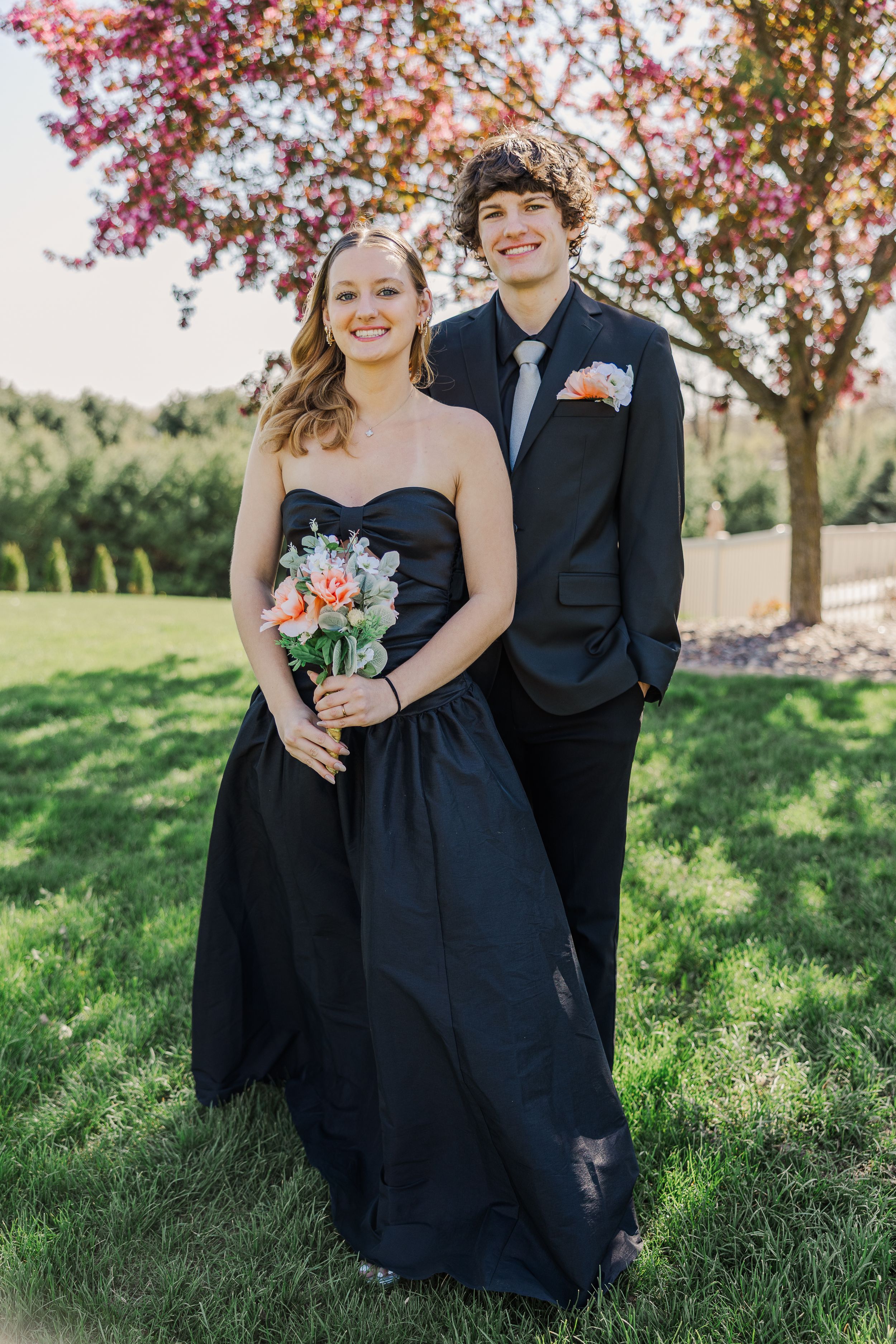 A couple in formal attire poses beneath a blooming cherry blossom tree.