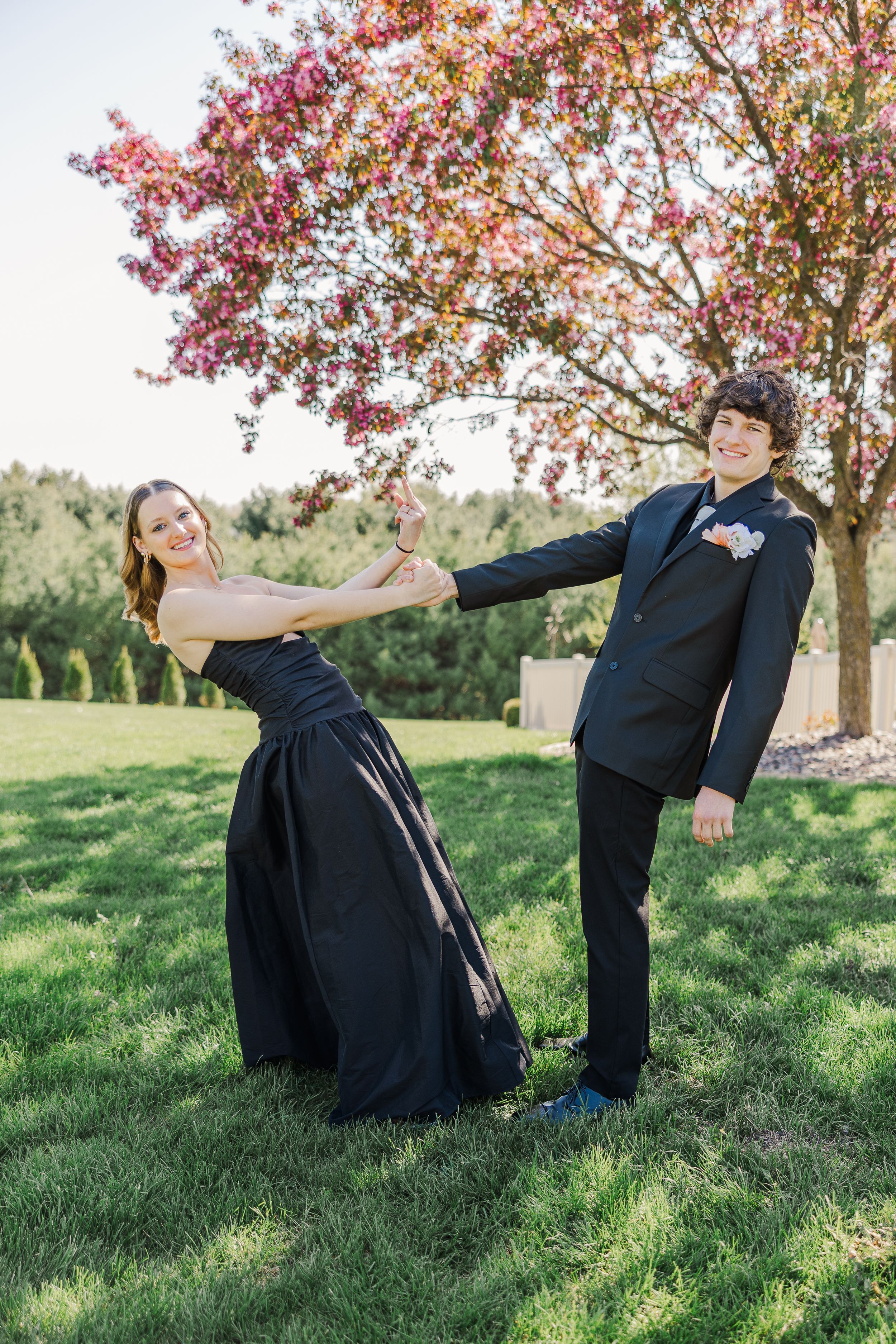 A couple in formal wear strikes a dance pose under flowering spring trees.