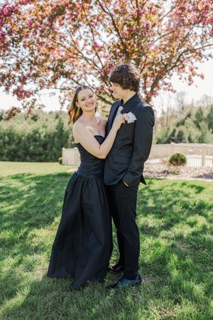 A couple in formal black attire poses playfully together on green grass under spring blossoms.