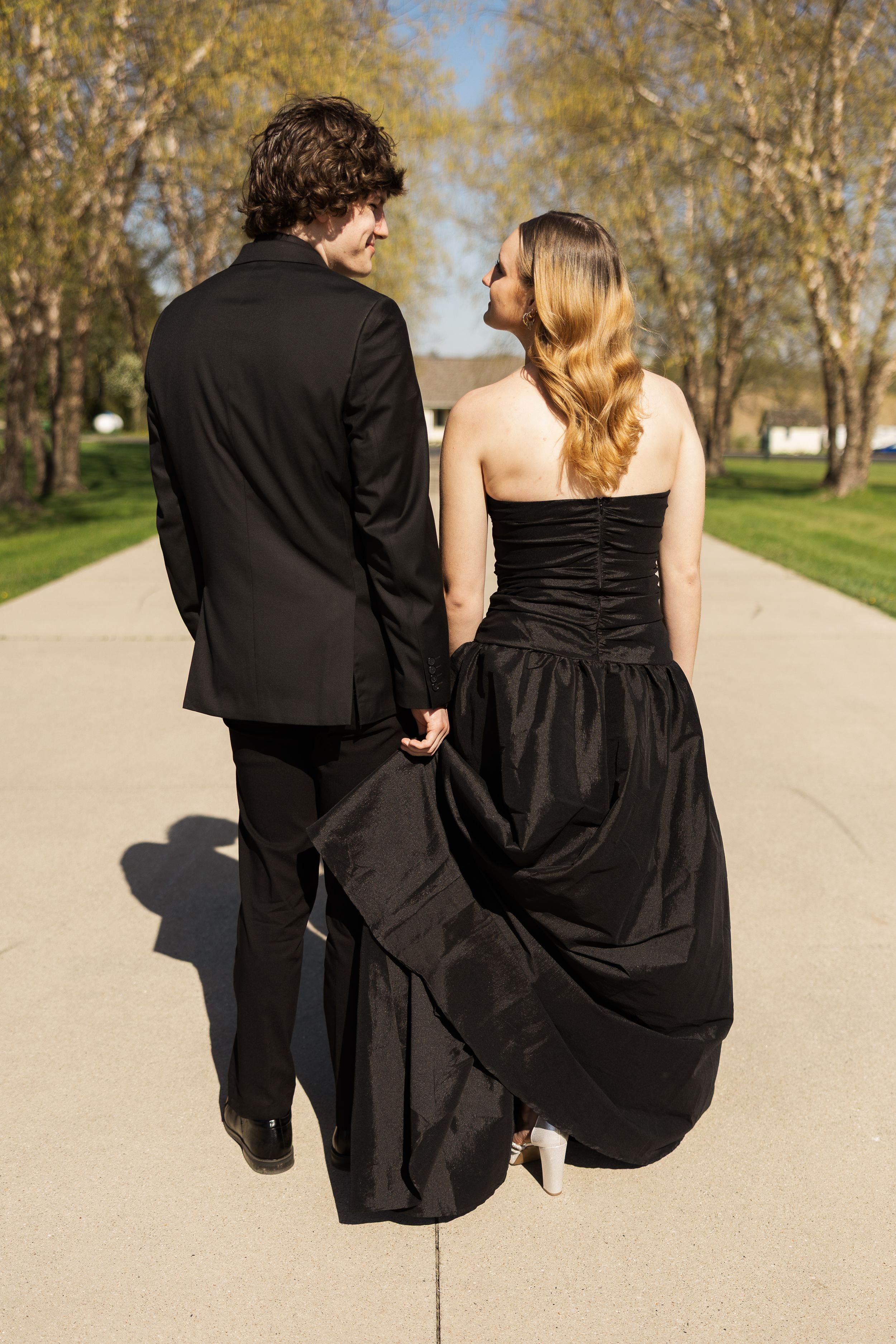 Couple in matching black formalwear walking together along a concrete pathway outdoors.