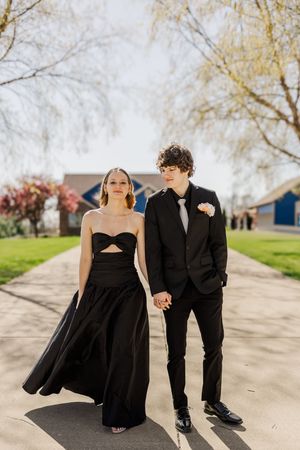 A couple dressed in black formal attire strolling together along a sidewalk on a spring day.