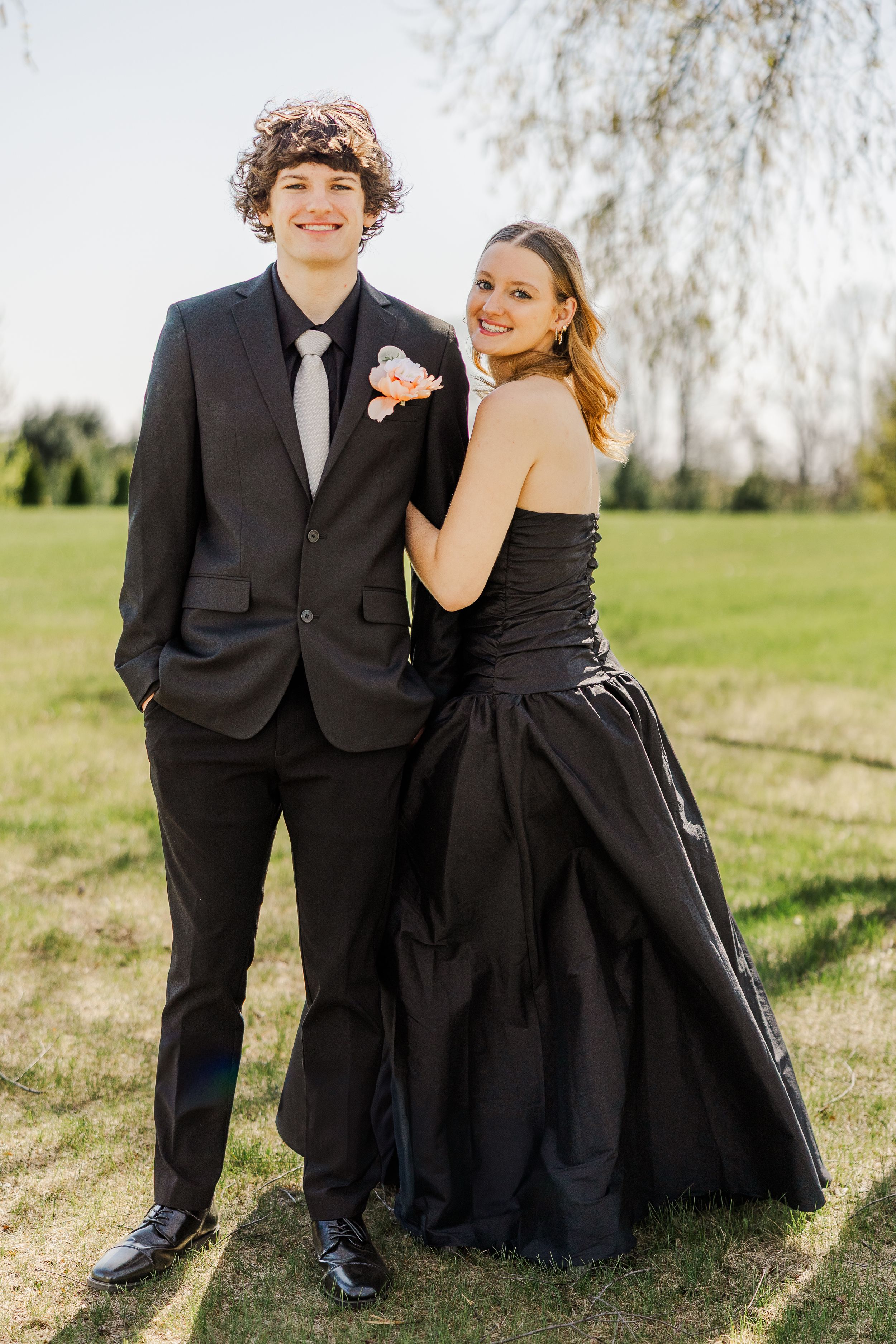Couple in black formal attire pose together on grassy hill with willow trees in background.