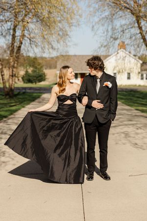 Couple in black formal attire dances and poses together on concrete path outdoors.