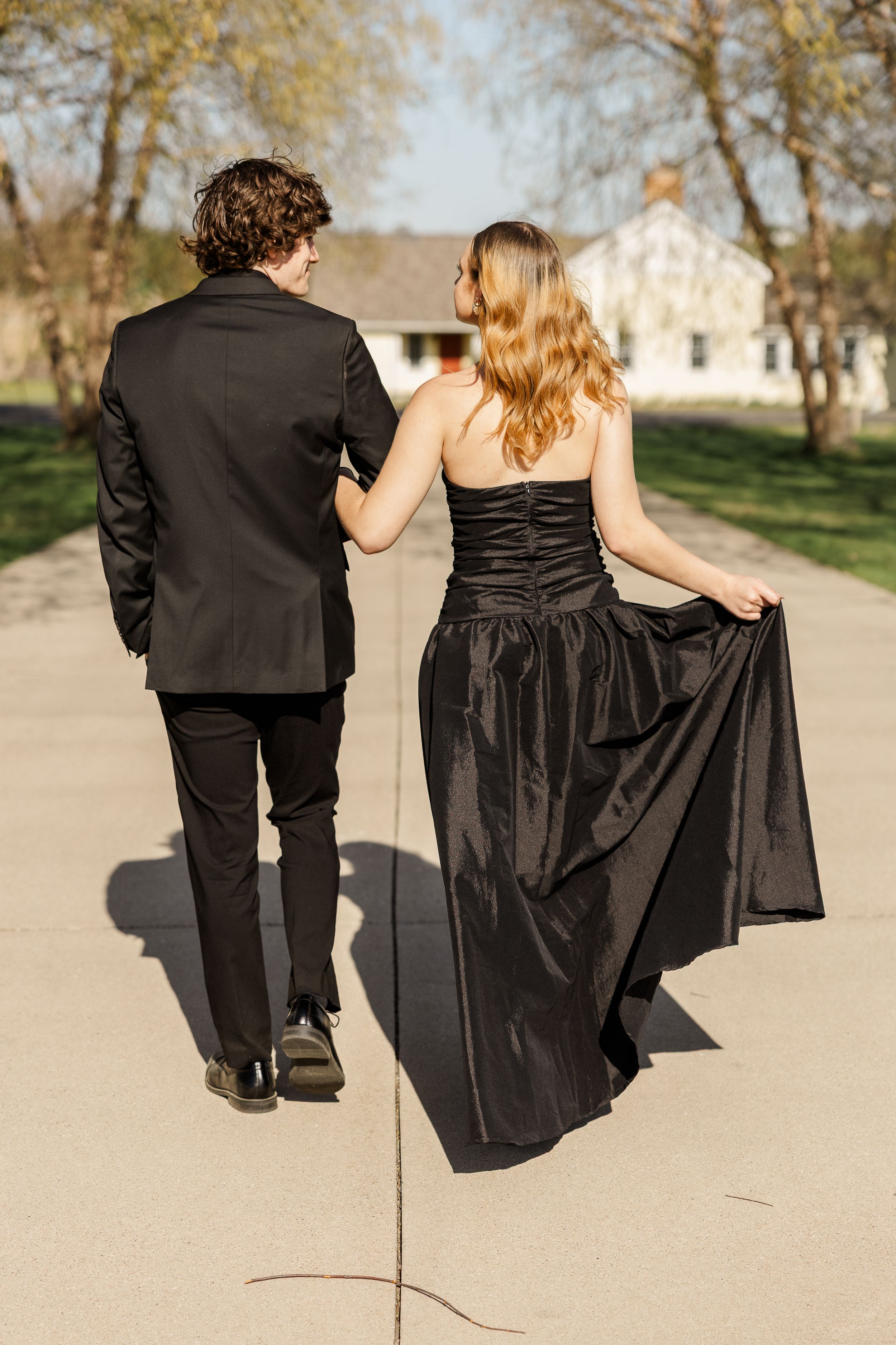 A couple in formal attire walking down a path, with the woman holding her black evening gown and a house visible in background.