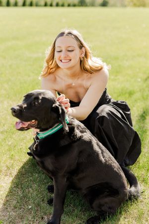 A black dog wearing a green collar sits on grass outdoors on a sunny day.