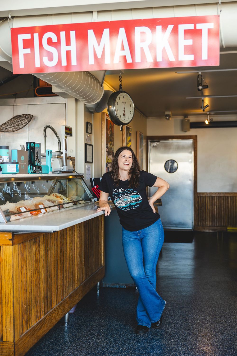 Staff member stands behind wooden counter at a fish market with red signage visible in background.