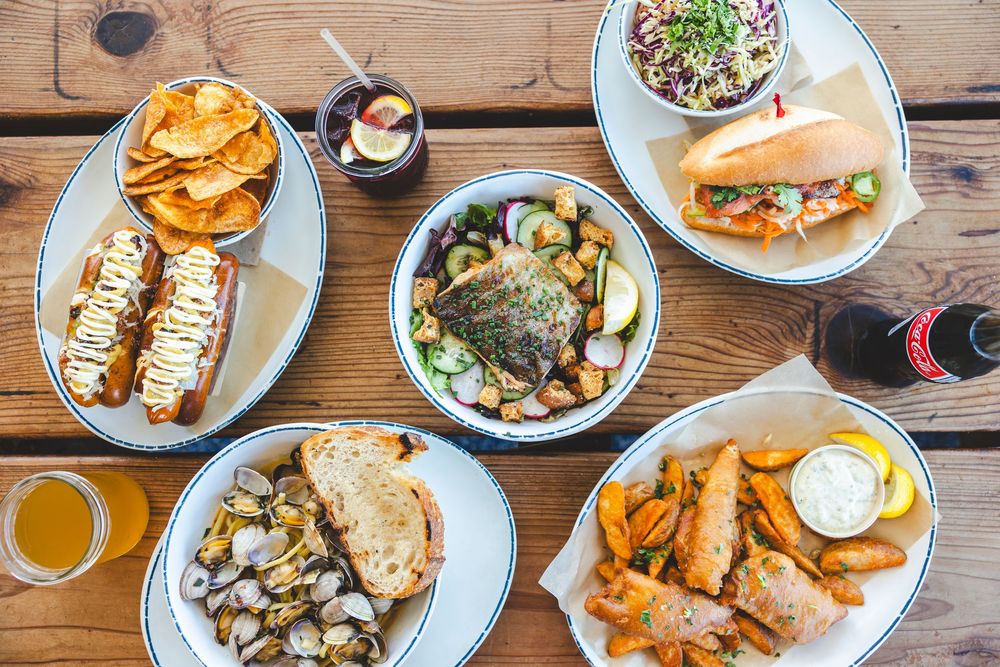 Overhead view of various seafood dishes and sandwiches served on wooden tables with drinks.