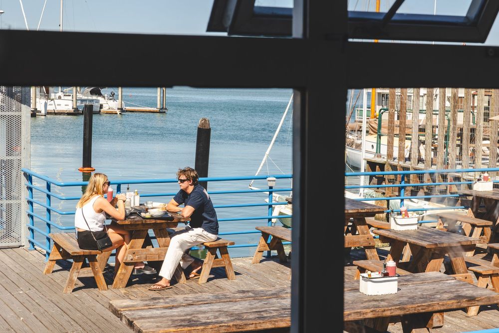 People dining at wooden picnic tables on a waterfront deck overlooking a marina on a sunny day.
