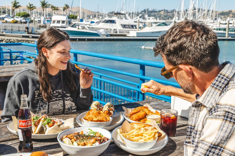 Diners enjoy waterfront dining at an outdoor restaurant with marina views and boats in the background.
