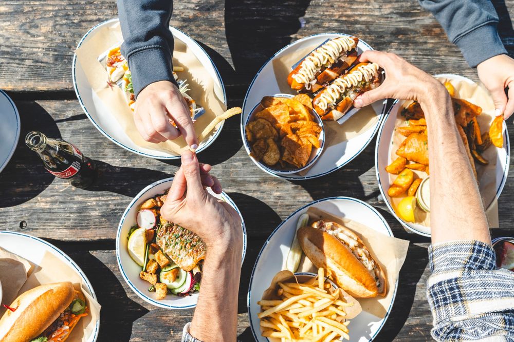 Hands reaching for burgers, fries and sandwiches on wooden table during casual dining.