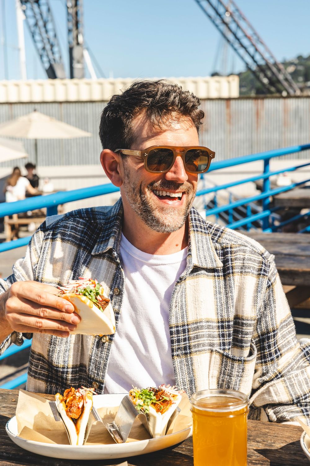 Person enjoying a casual outdoor meal with beer and sandwiches at a sunny waterfront restaurant.