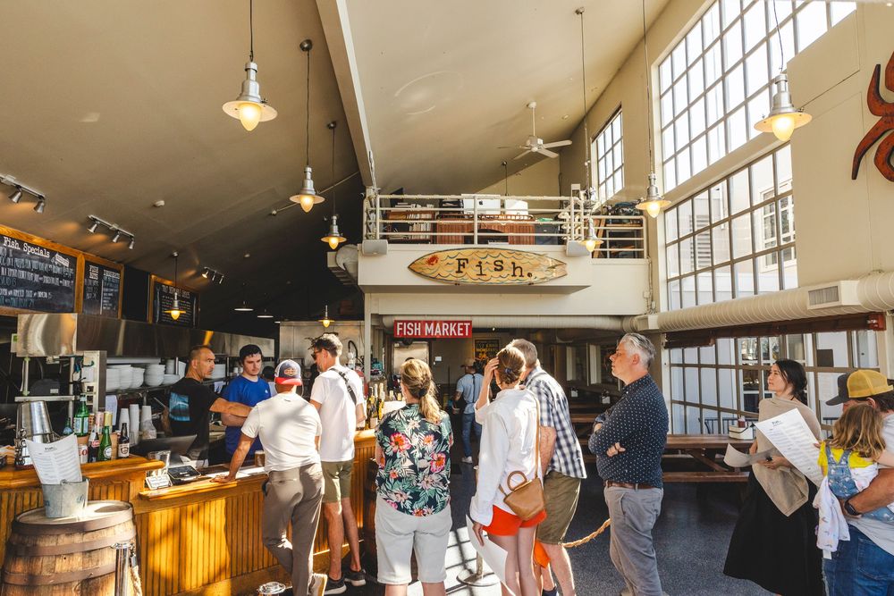 Customers line up inside a bustling marketplace with high ceilings, pendant lights and large windows.