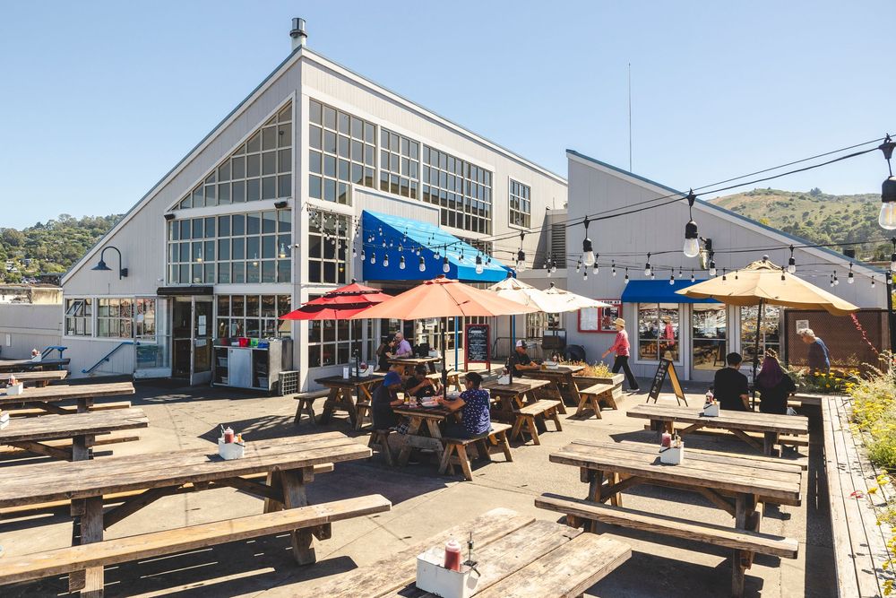 Outdoor dining area with wooden picnic tables and umbrellas in front of a modern waterfront building with glass windows.