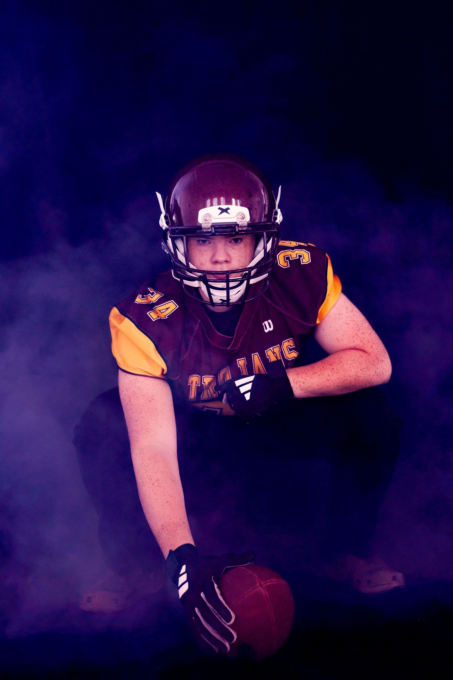 American football player in maroon and gold uniform holding ball, surrounded by dramatic purple smoke on dark background.
