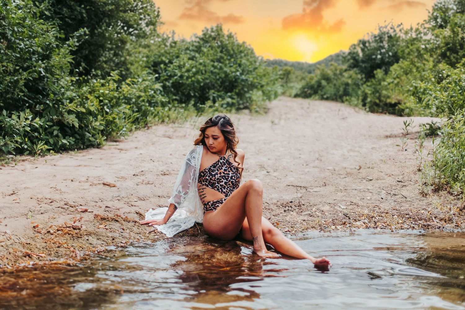 A person in floral swimwear sits at the edge of a stream at sunset with lush greenery surrounding the dirt path.