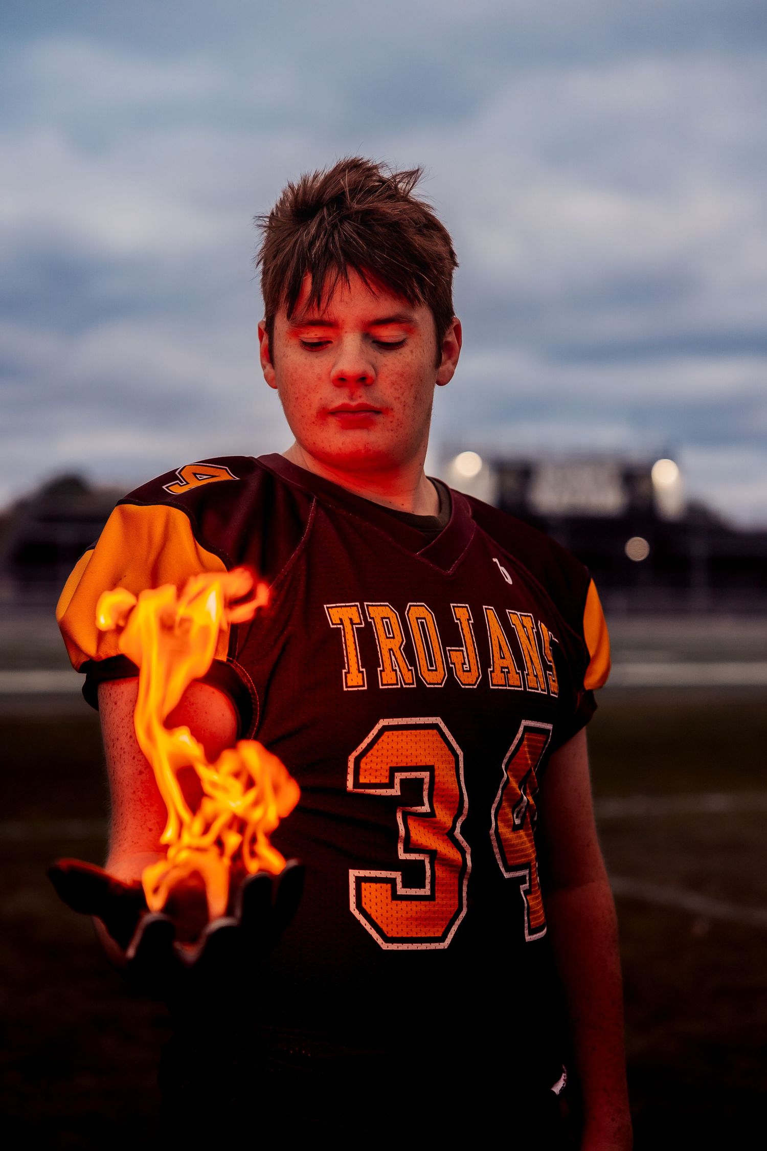 A Trojans football player wearing jersey number 34 stands on a field with flames in his hand at dusk.