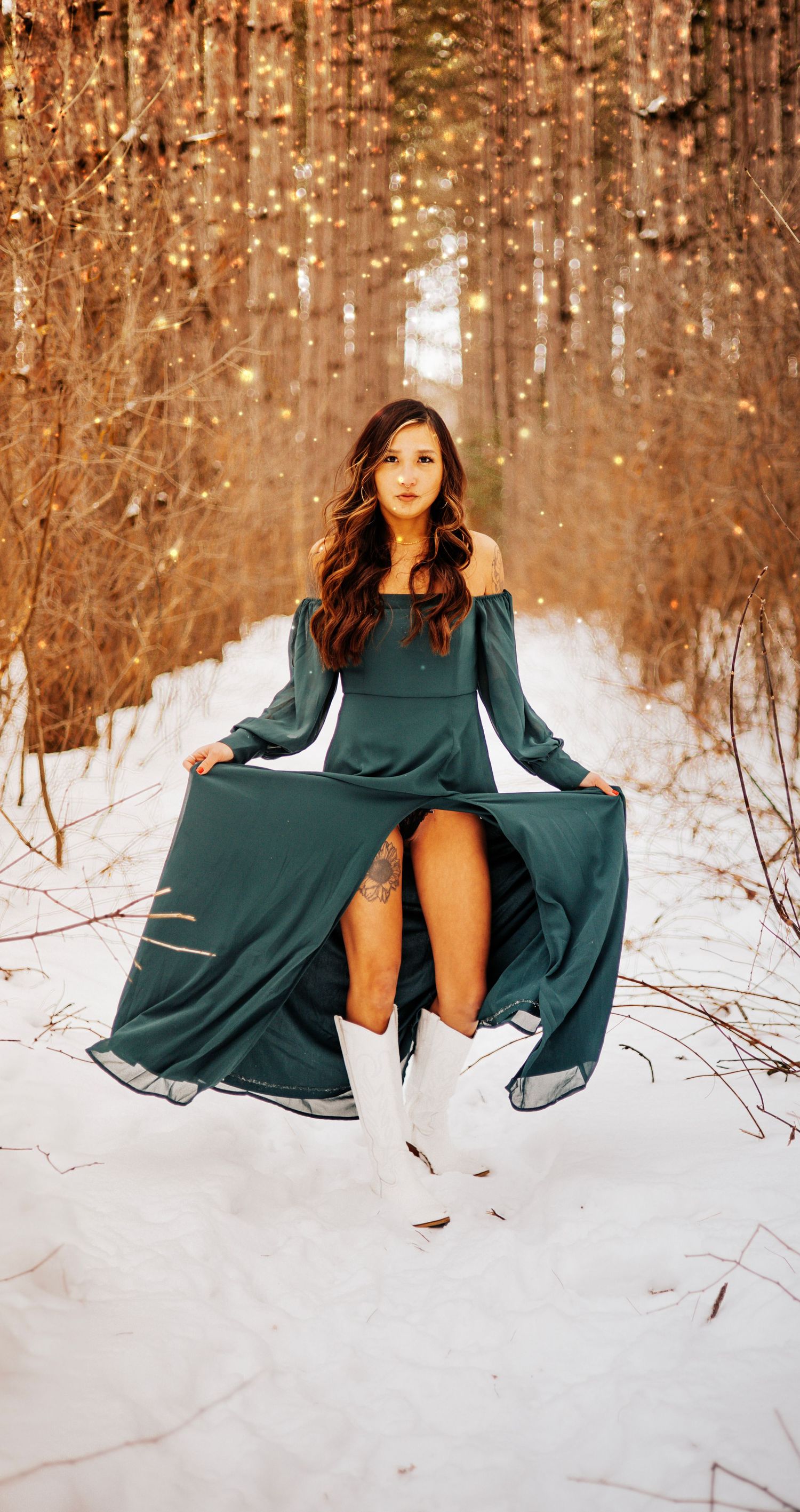 A person in a flowing emerald green dress poses gracefully in a snowy forest with warm autumn-colored trees.