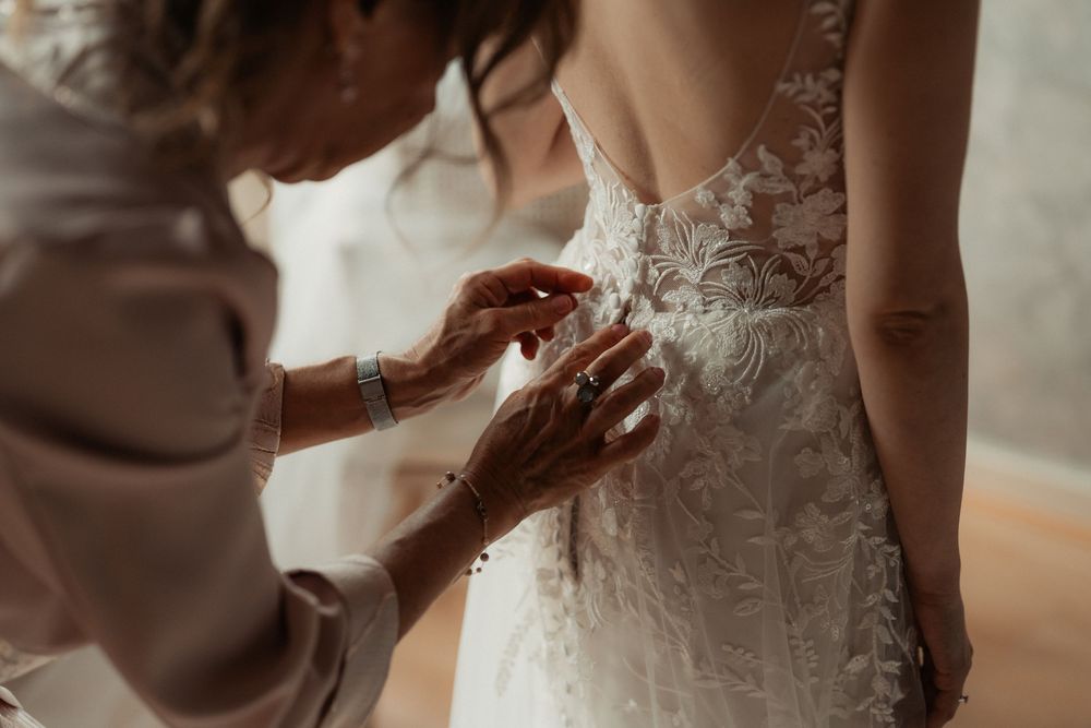 Someone helps adjust the intricate lace details on a white wedding dress during a bridal preparation moment.