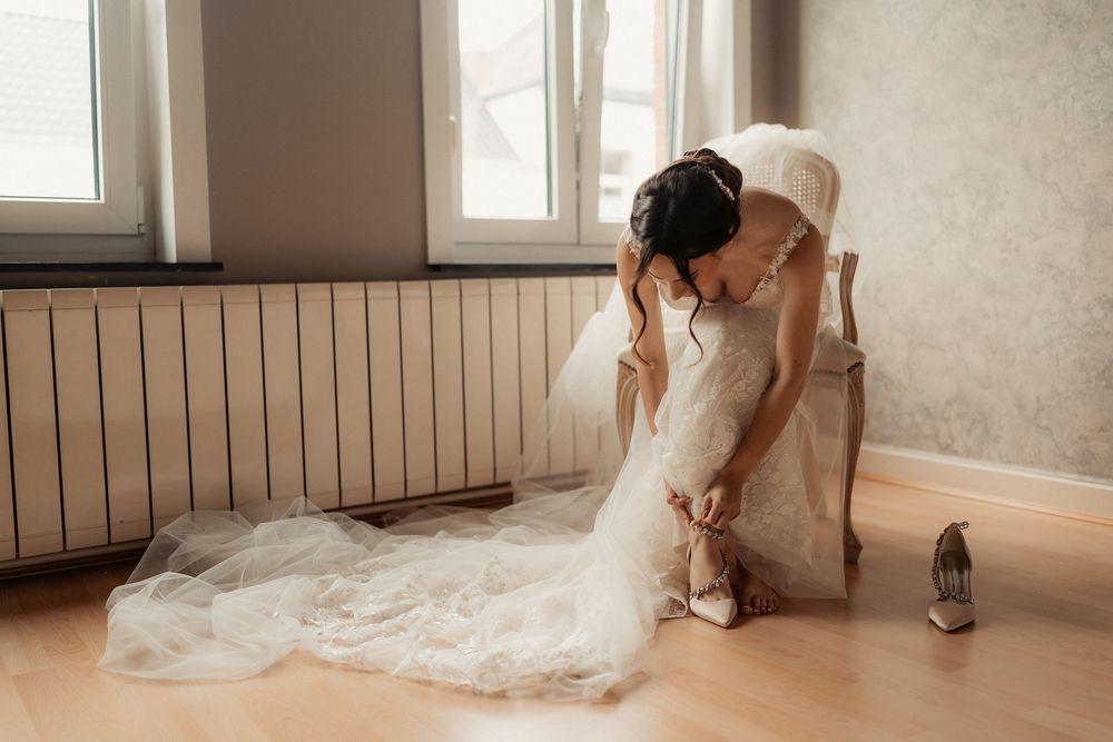 A bride in a flowing white lace gown adjusts her shoes while getting ready in a sunlit room.