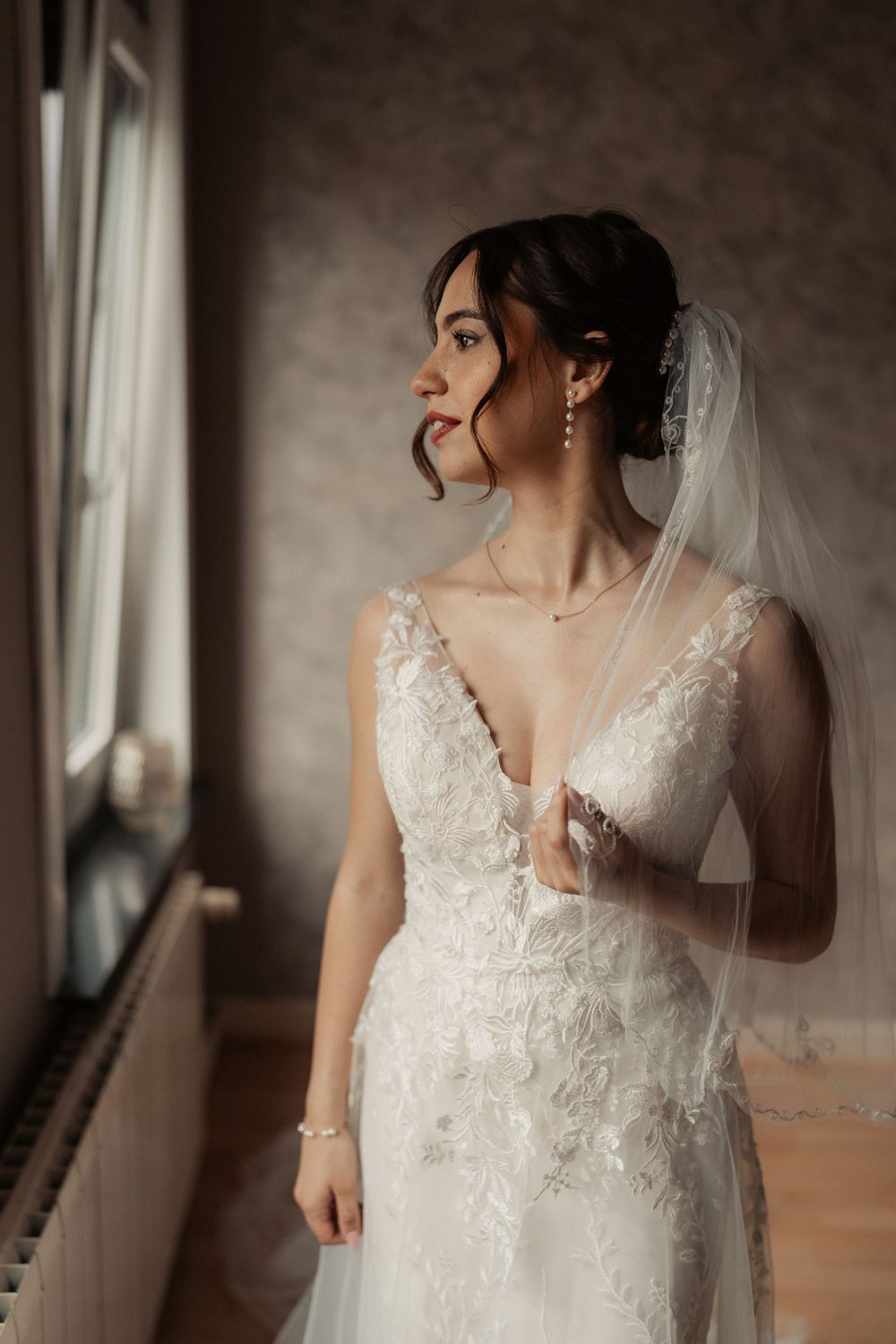 A bride in an elegant white lace wedding dress and veil gazes thoughtfully by a window in moody lighting.